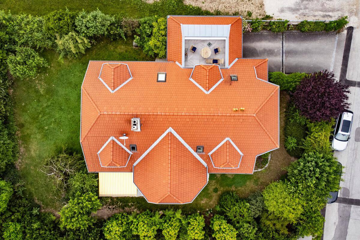 Vogelperspektive des Hauses mit rotem Ziegeldach und einer kleinen Dachterrasse, umgeben von Bäumen.