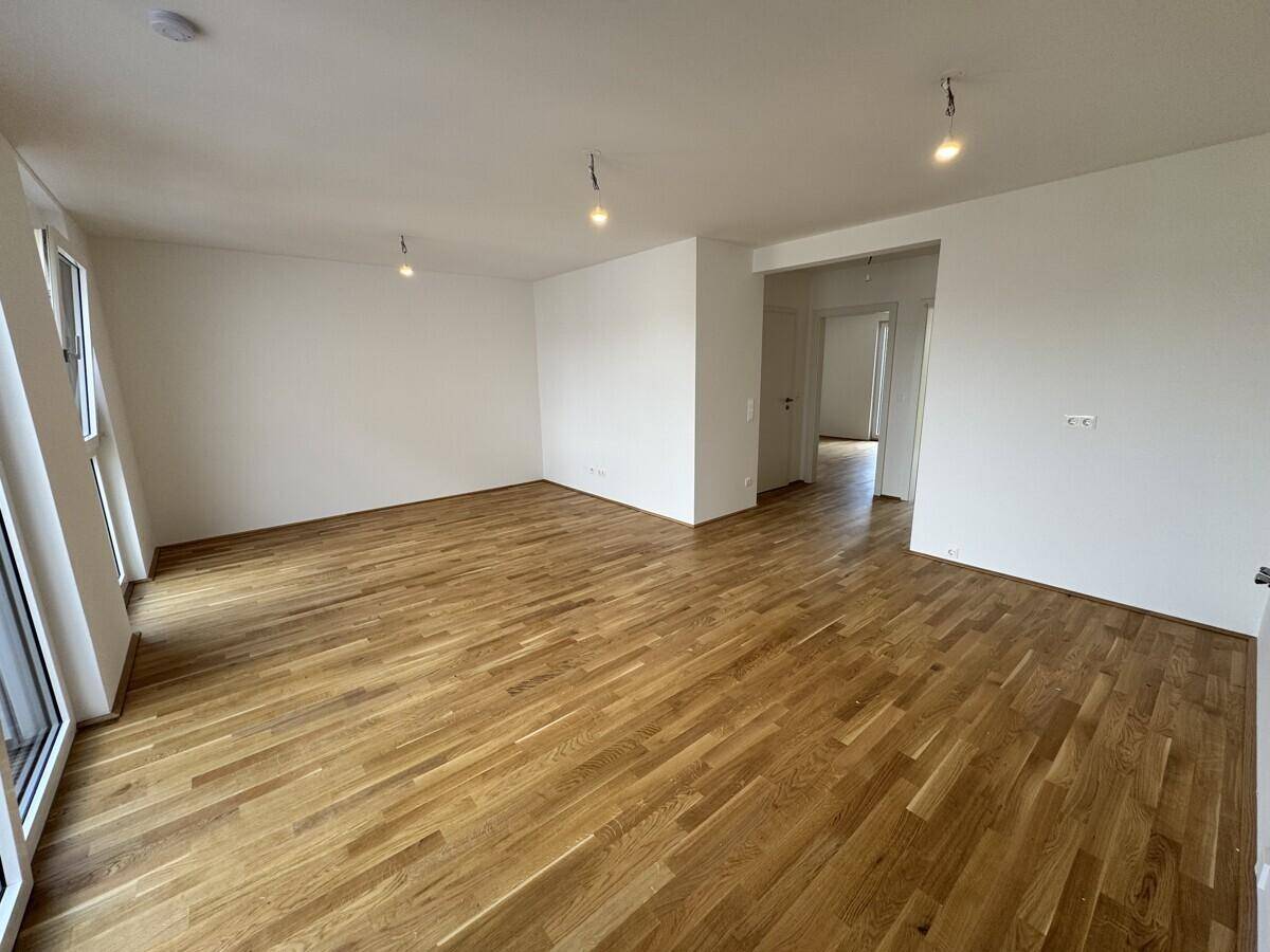 Helles, leeres Wohnzimmer mit neuem Parkettboden und großen Fenstern für viel Tageslicht.