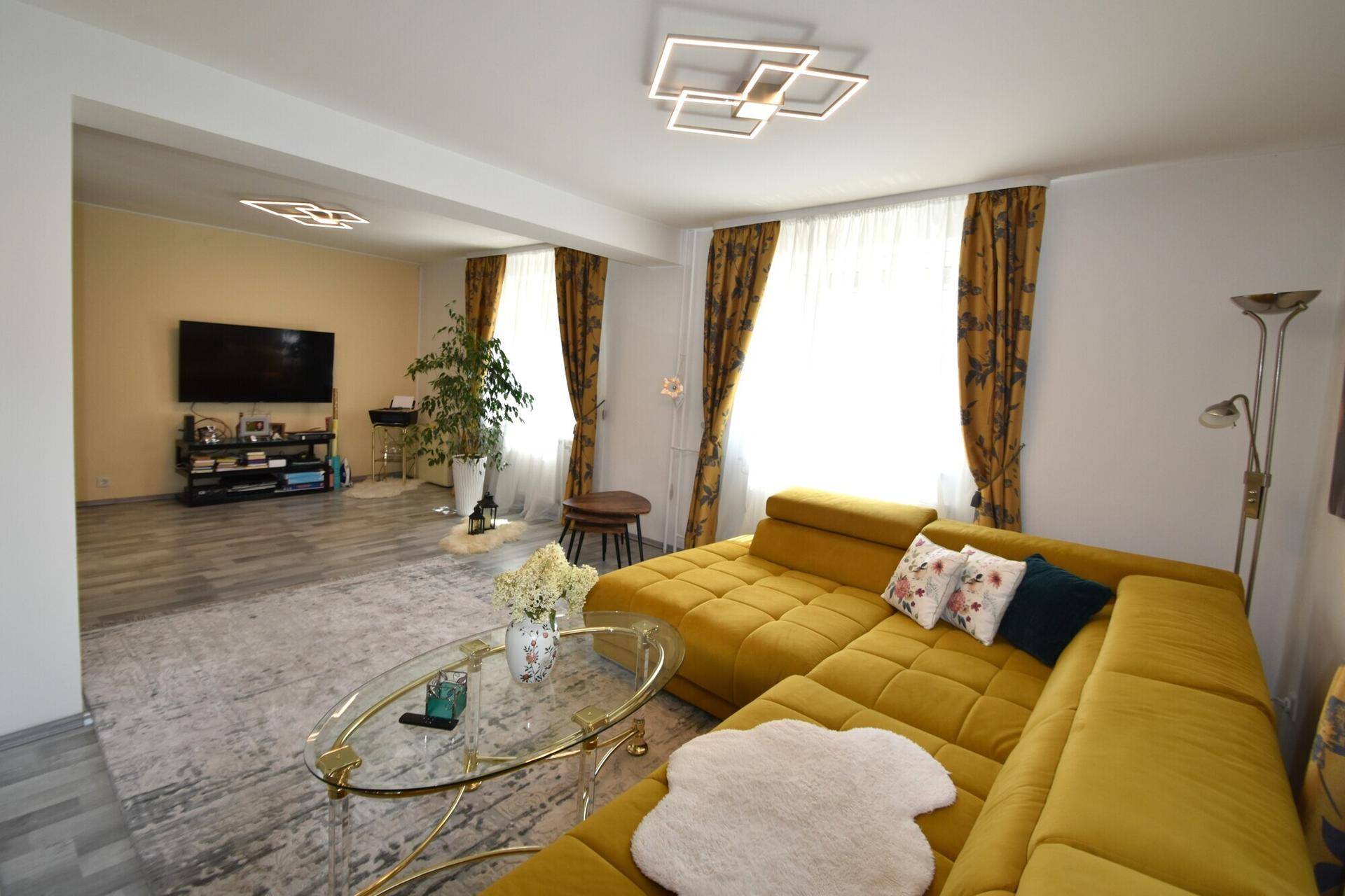 Geräumiges Wohnzimmer mit gelbem Ecksofa, Fernseher und modernem Deckenlicht.