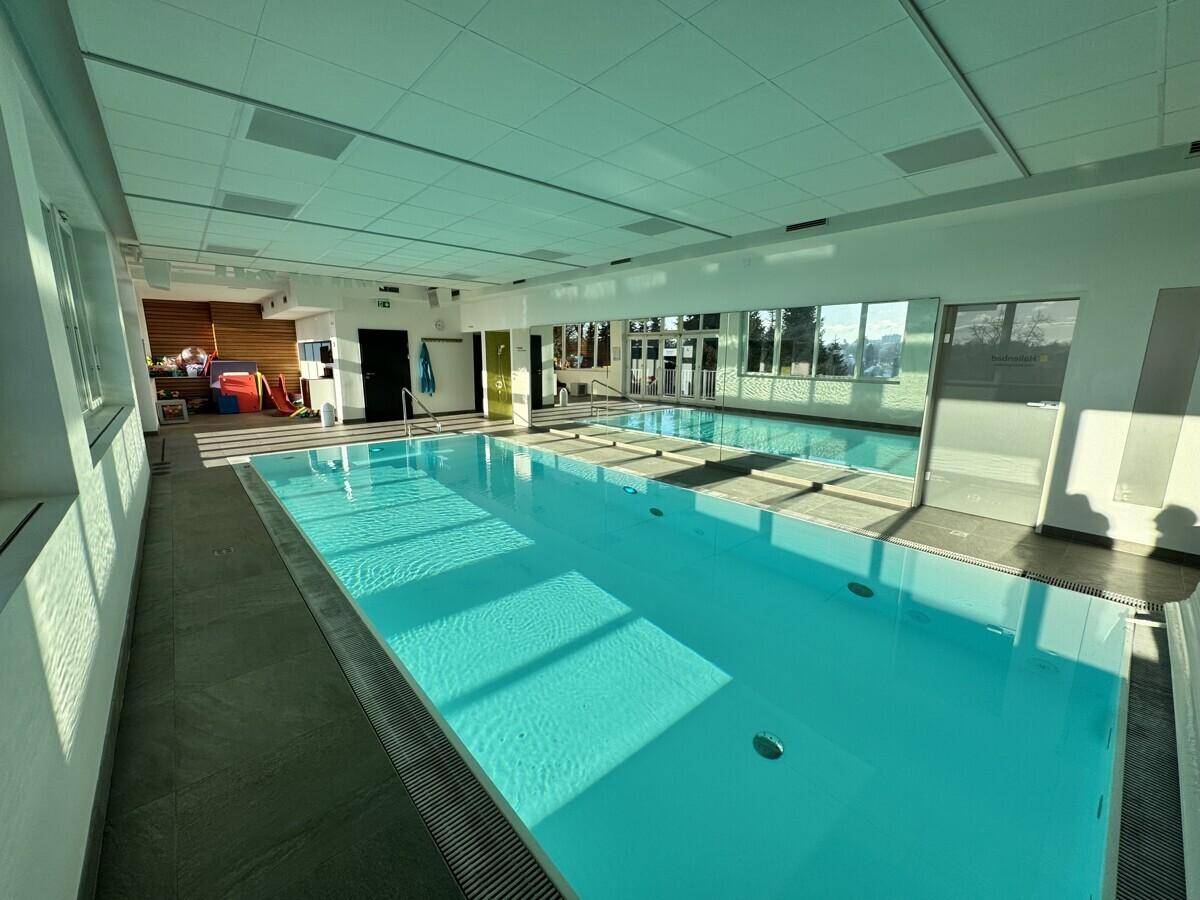 Großzügiges Hallenbad mit hellblauem Wasser, großen Fenstern und Zugang zu einem Außenbereich.