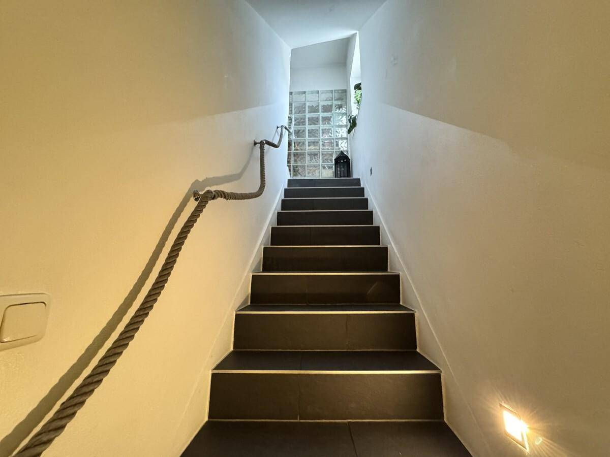 Schmale Treppe mit dunklen Stufen und einem rustikalen Seilgeländer, beleuchtet von Wandleuchten.