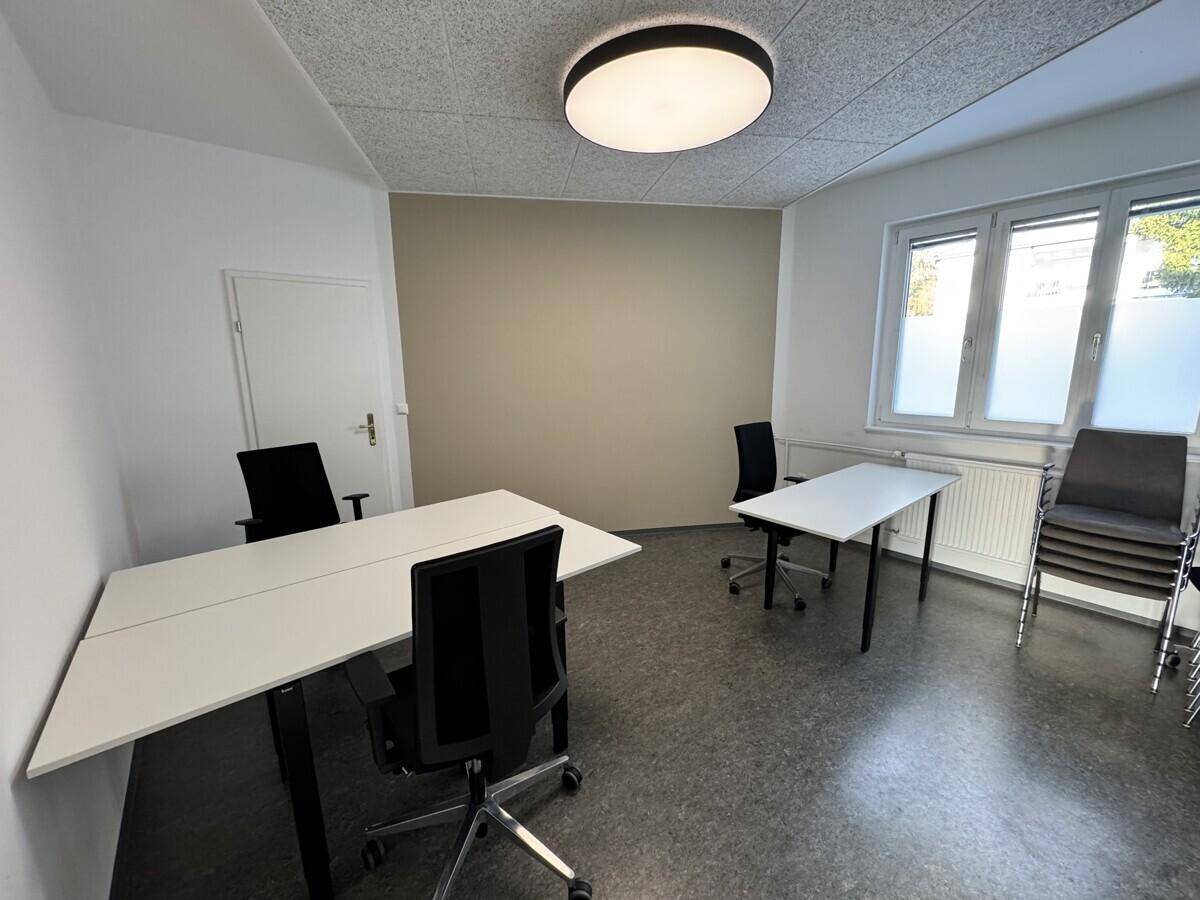 Heller Büroraum mit mehreren Schreibtischen, Bürostühlen und großen Fenstern, die viel Licht hereinlassen.