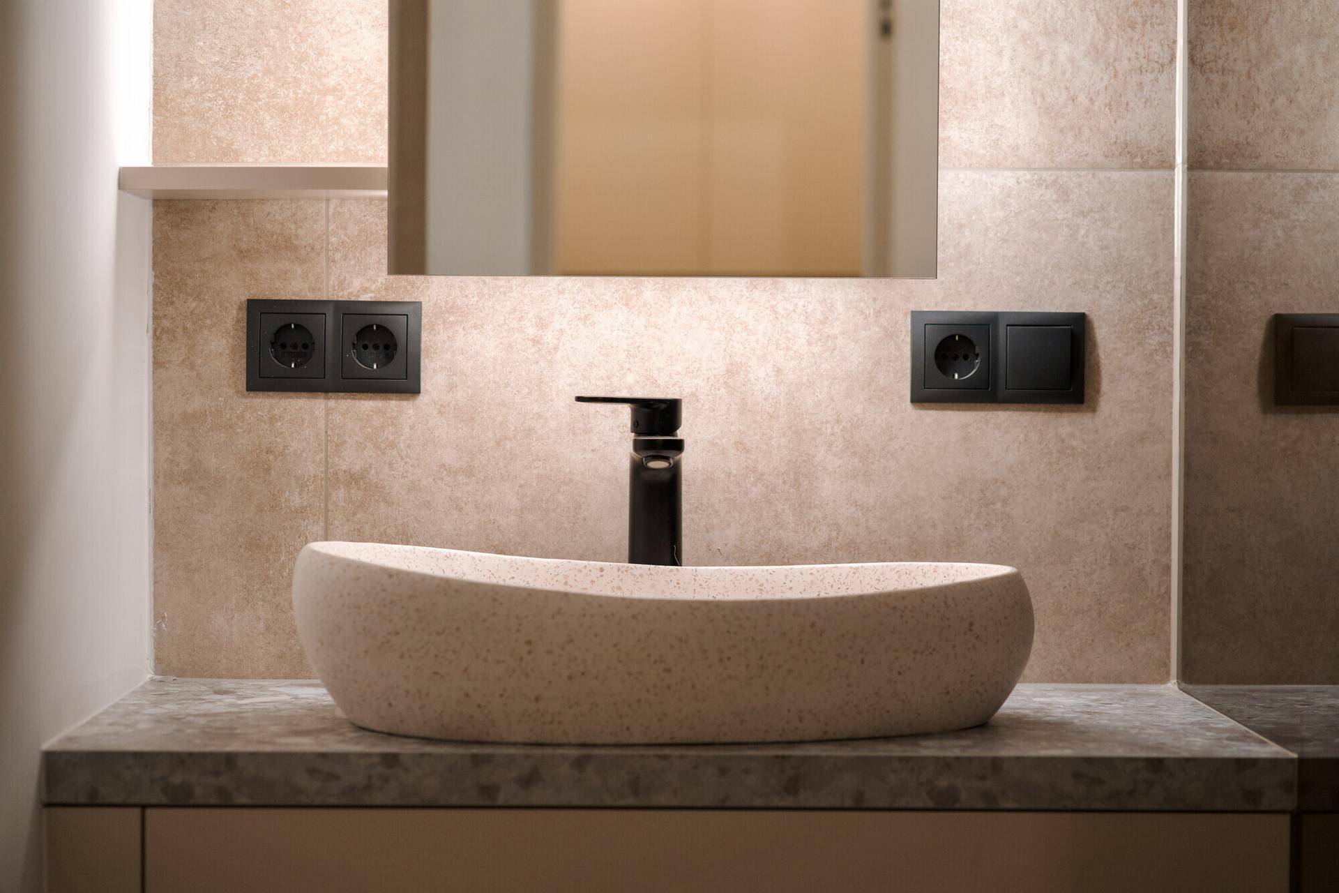 Modernes Badezimmer mit beigefarbenen Fliesen, ovalem Aufsatzwaschbecken und schwarzer Armatur.
