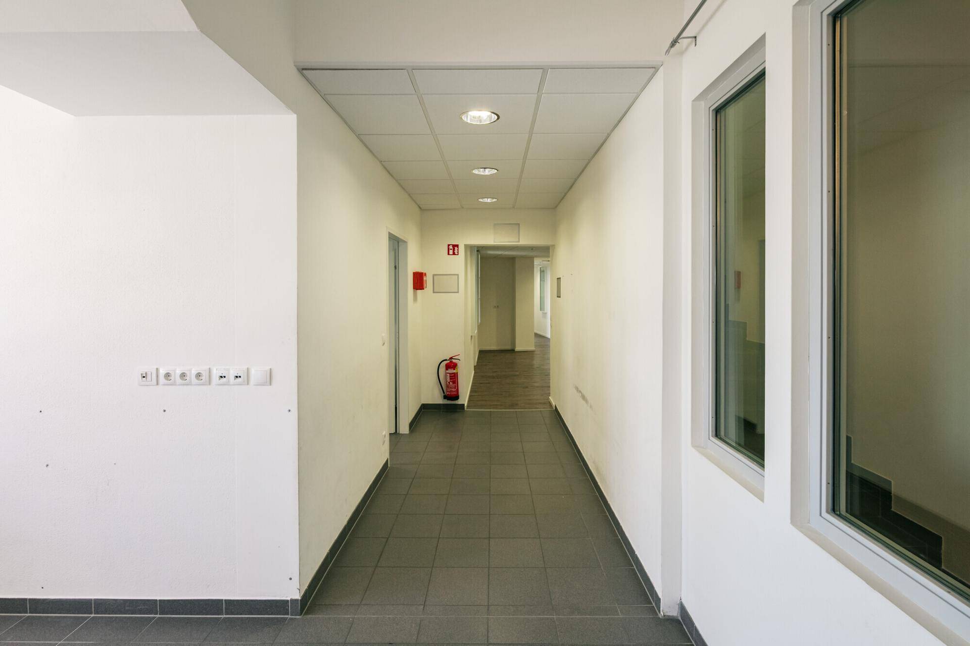 Langer Flur mit dunklen Fliesen und weißen Wänden, der zu weiteren Räumen und einem Treppenhaus führt.