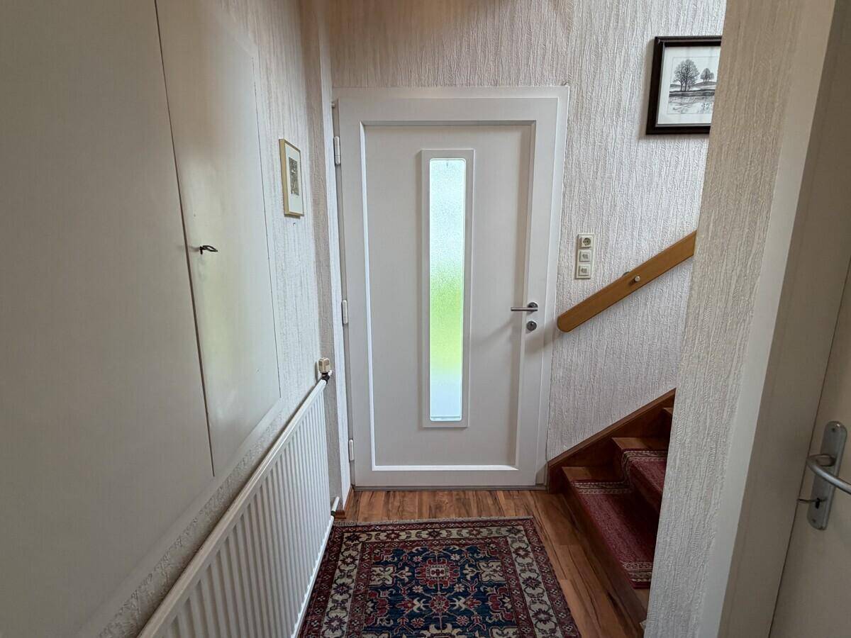 Eingangsbereich mit heller Tür, Holztreppe und Teppichläufer, der ins Obergeschoss führt.