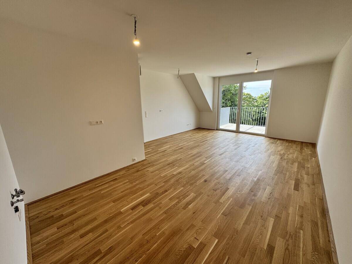 Helles, leeres Zimmer mit Holzboden und Zugang zu einem kleinen Balkon mit Blick ins Grüne.