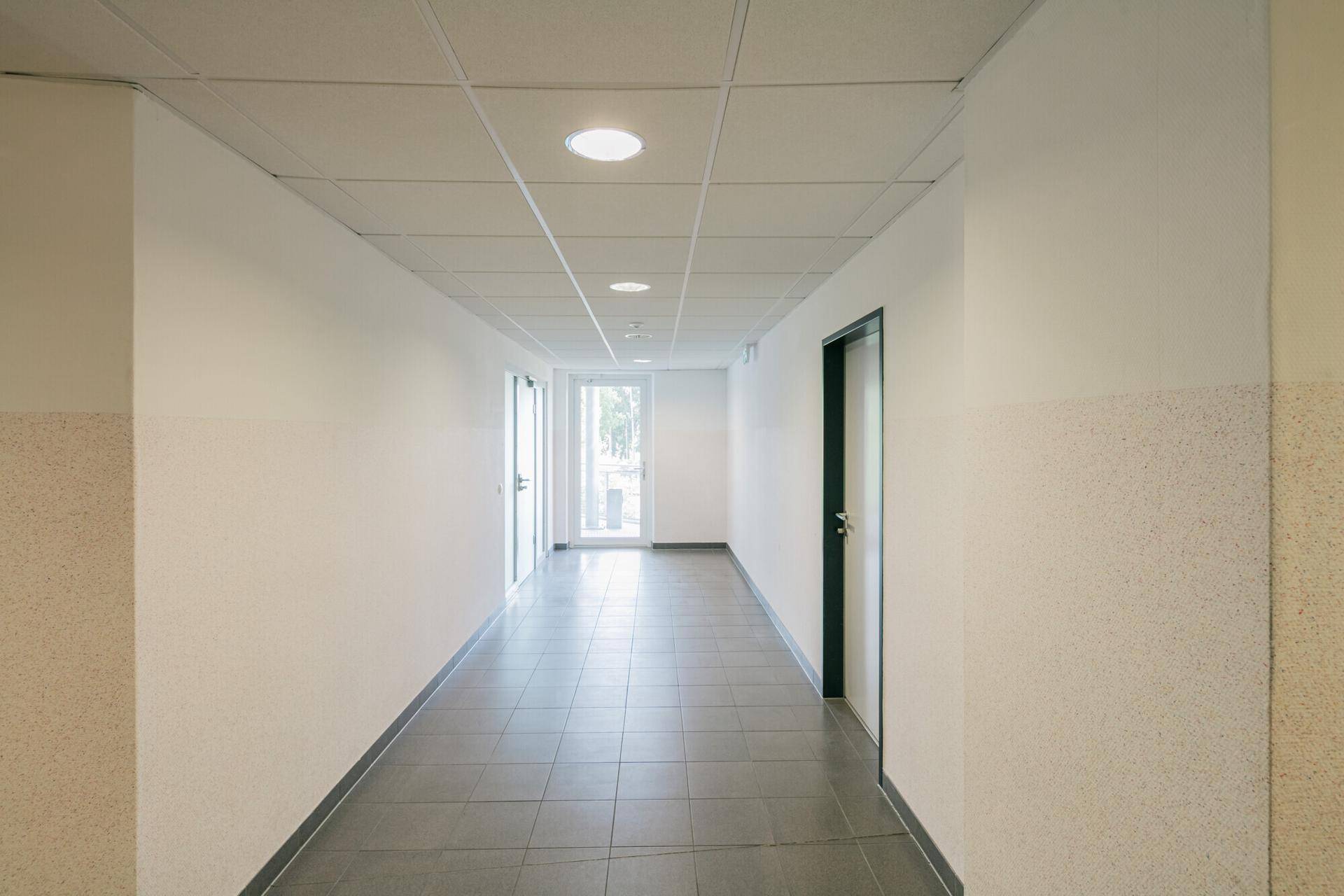 Langer, heller Flur mit grauen Fliesen und abgehängter Decke, führt zu weiteren Räumen.