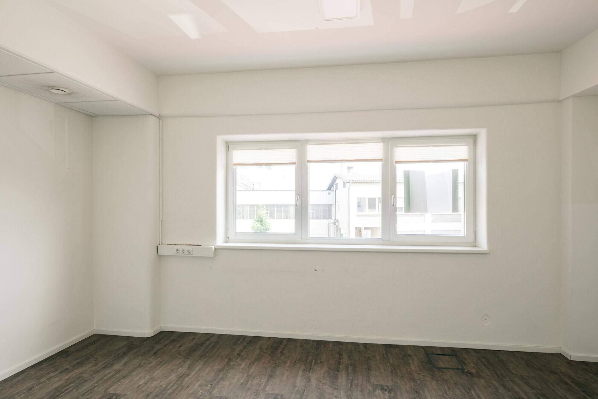 Leerer Büroraum mit großen Fenstern, die viel Tageslicht hereinlassen, und Holzoptik-Boden.