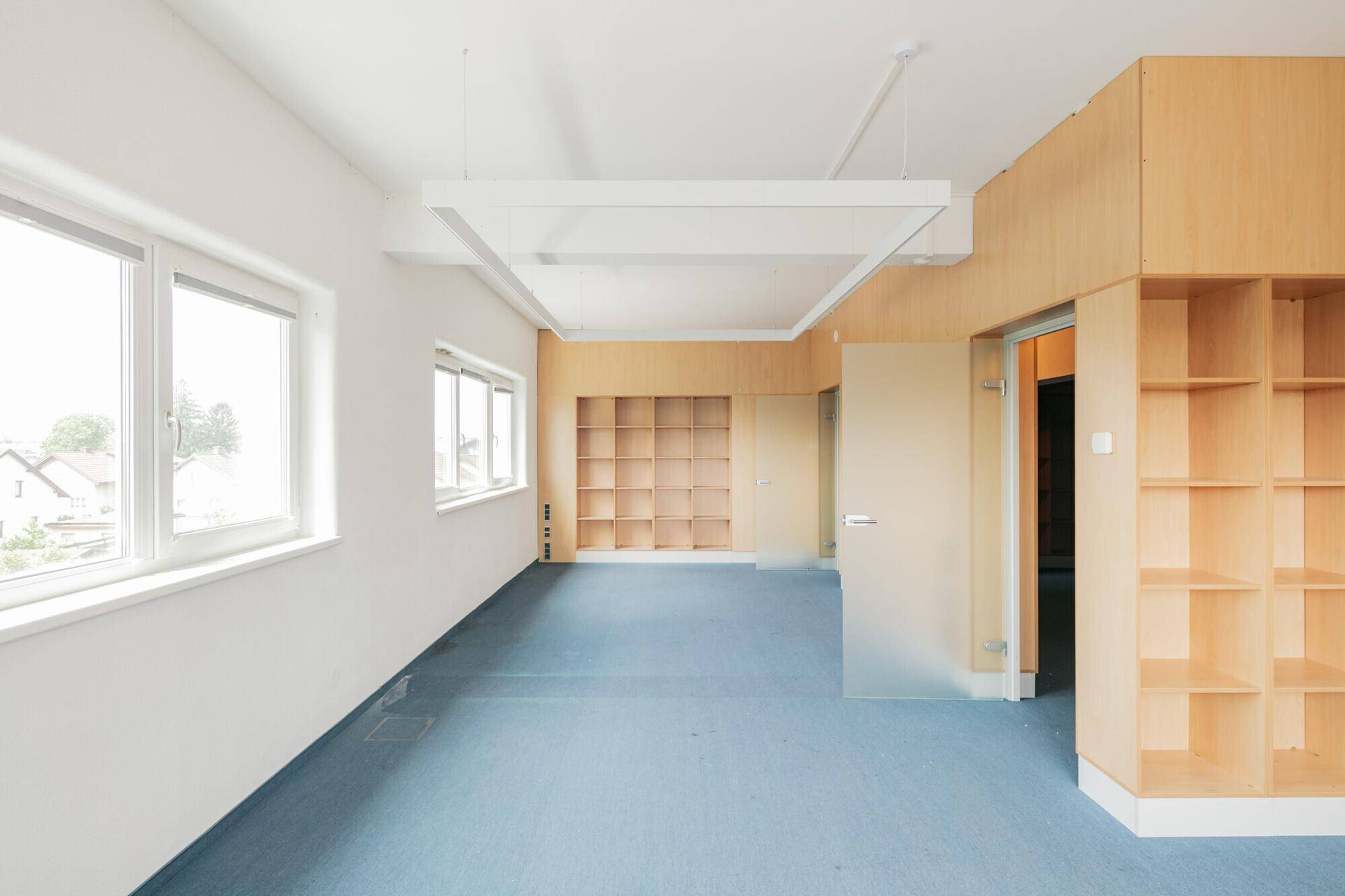 Helles Büro mit blauem Teppichboden, Holzverkleidungen, integrierten Regalen und einer modernen Glastür.