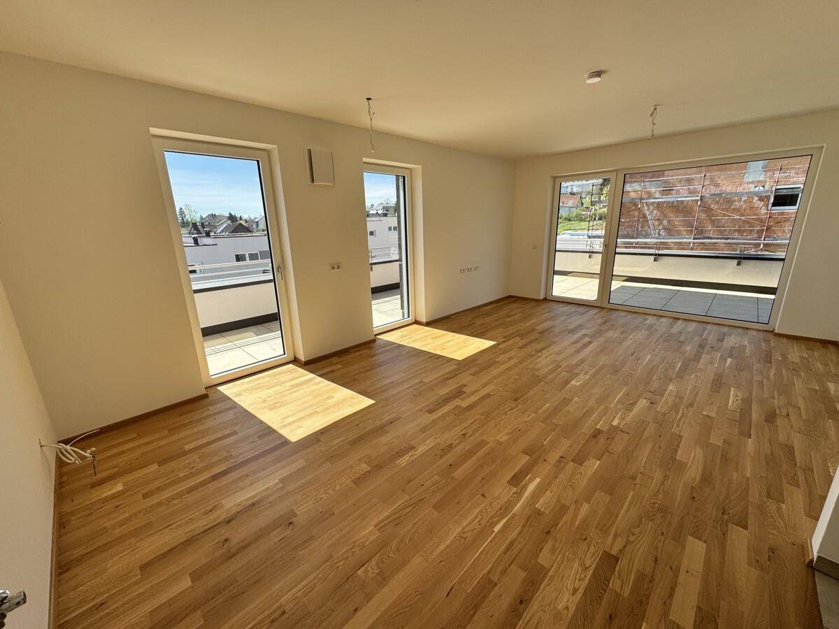 Heller, leerer Raum mit Holzboden und großen Fenstern, die auf eine Terrasse führen.