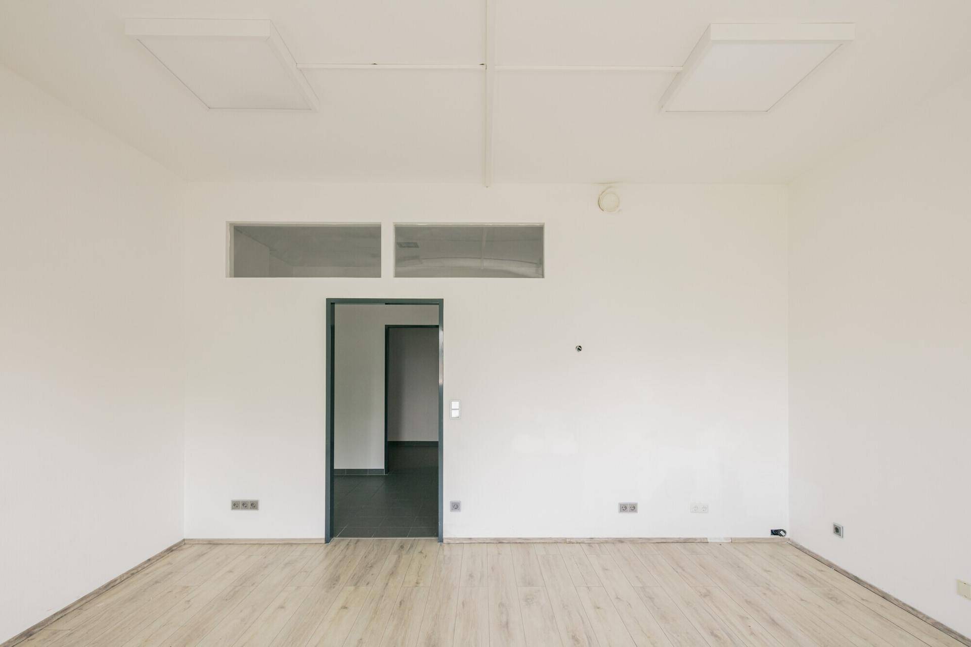 Leerer Büroraum mit hellem Laminatboden, weißen Wänden und Deckenleuchten.