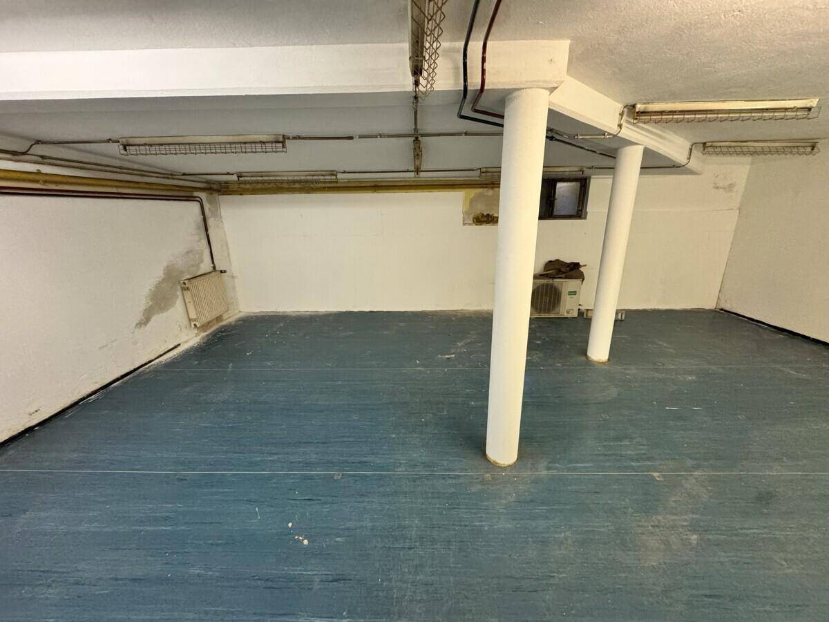 Kellerraum mit zwei tragenden Säulen, Heizkörper und einer kleinen Öffnung in der Wand, blaugrauer Boden.