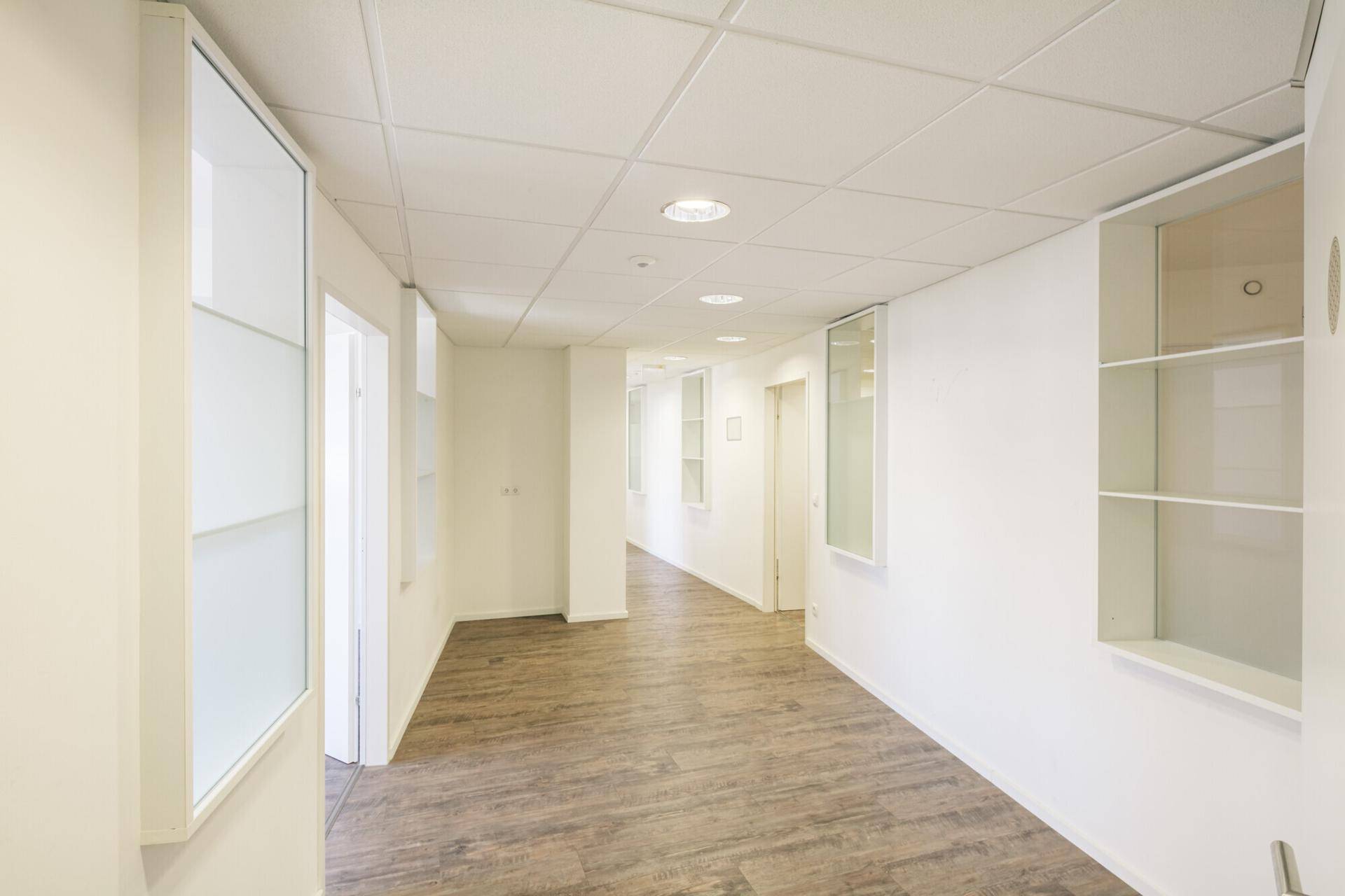 Langer, heller Flur mit Holzoptik-Boden und Einbauregalen, der zu mehreren Büroräumen führt.