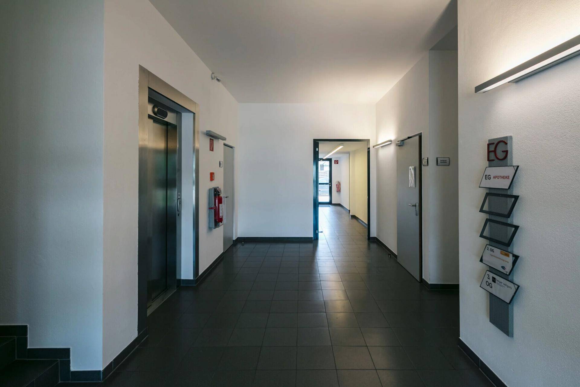 Langer, heller Flur mit Aufzug, dunklen Fliesen und Informationsschildern an der Wand.