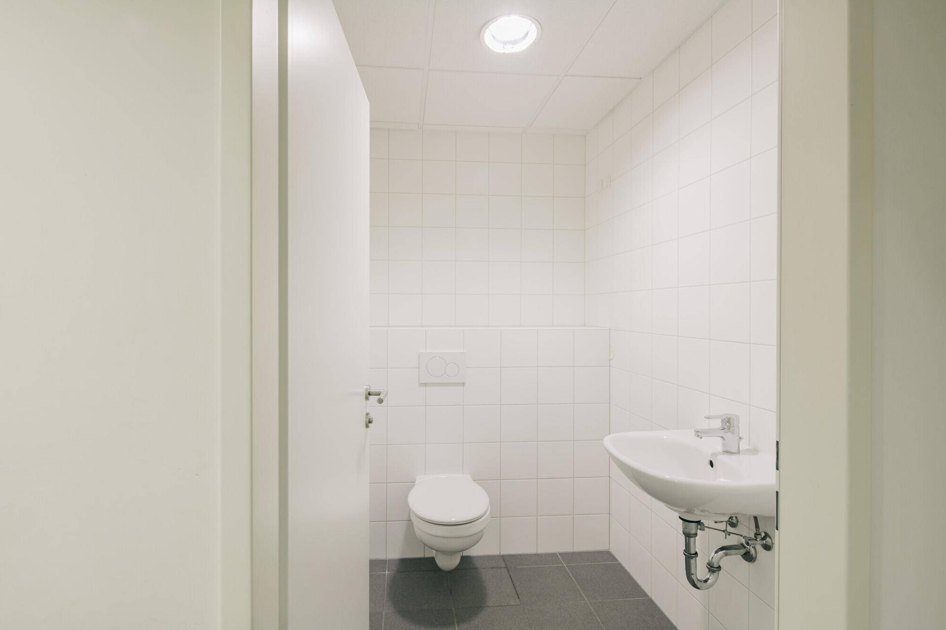 Saubere und funktionale Toilette mit weißen Fliesen, Wand-WC und Waschbecken in einem modernen Gebäude.