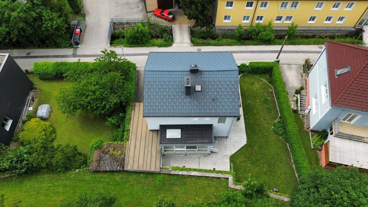 Draufsicht auf das Haus mit grauem Dach, umgeben von viel Grün und einer Holzterrasse im hinteren Bereich.