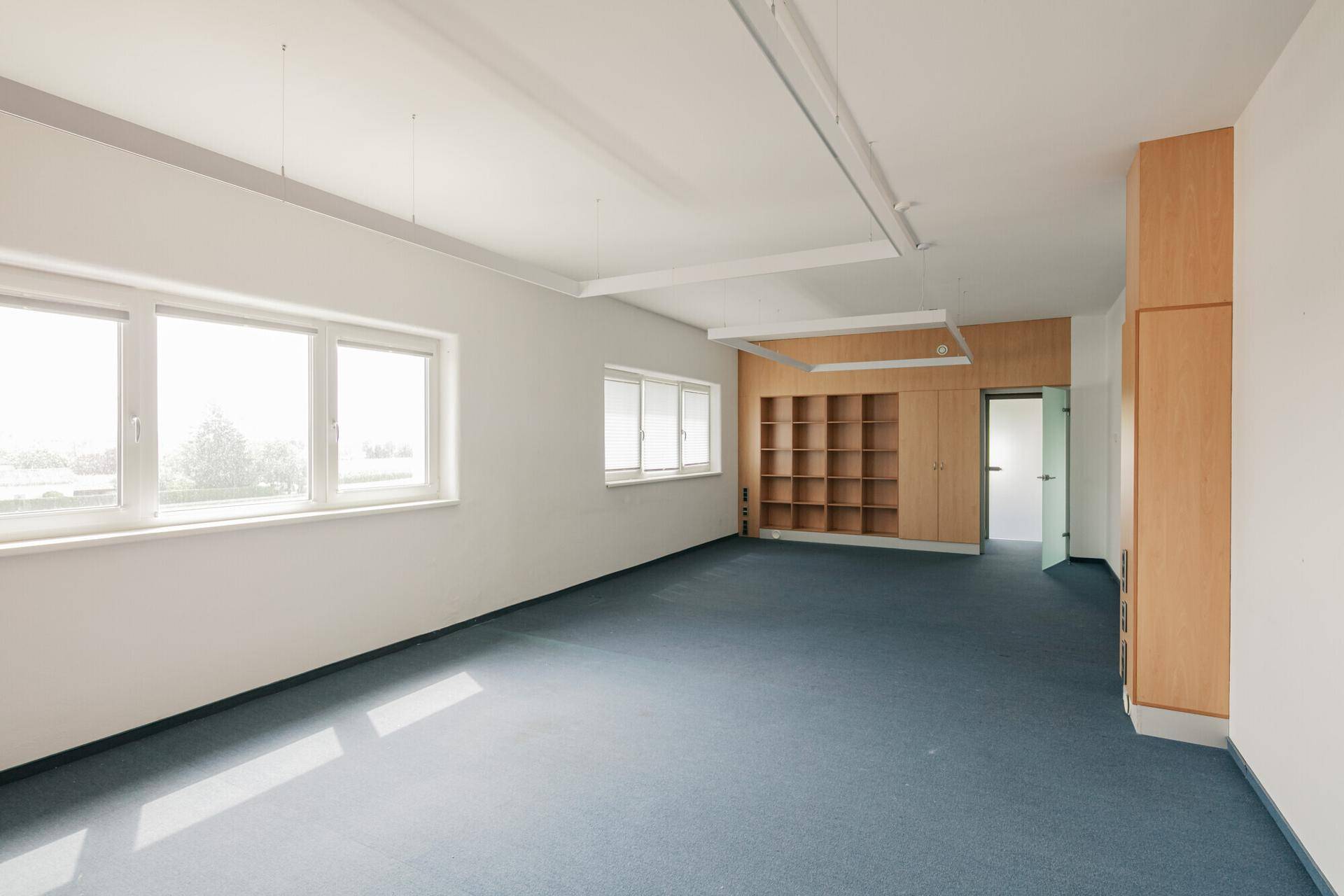 Langes Büro mit blauem Teppichboden, hellen Wänden und großen Fenstern, die viel Tageslicht hereinlassen.
