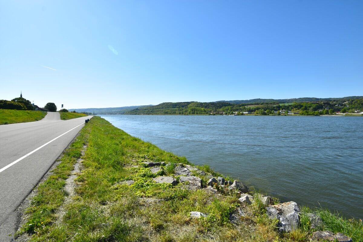 Malerischer Flusslauf mit Uferweg und bewaldeten Hügeln unter klarem Himmel, ideal für Spaziergänge.