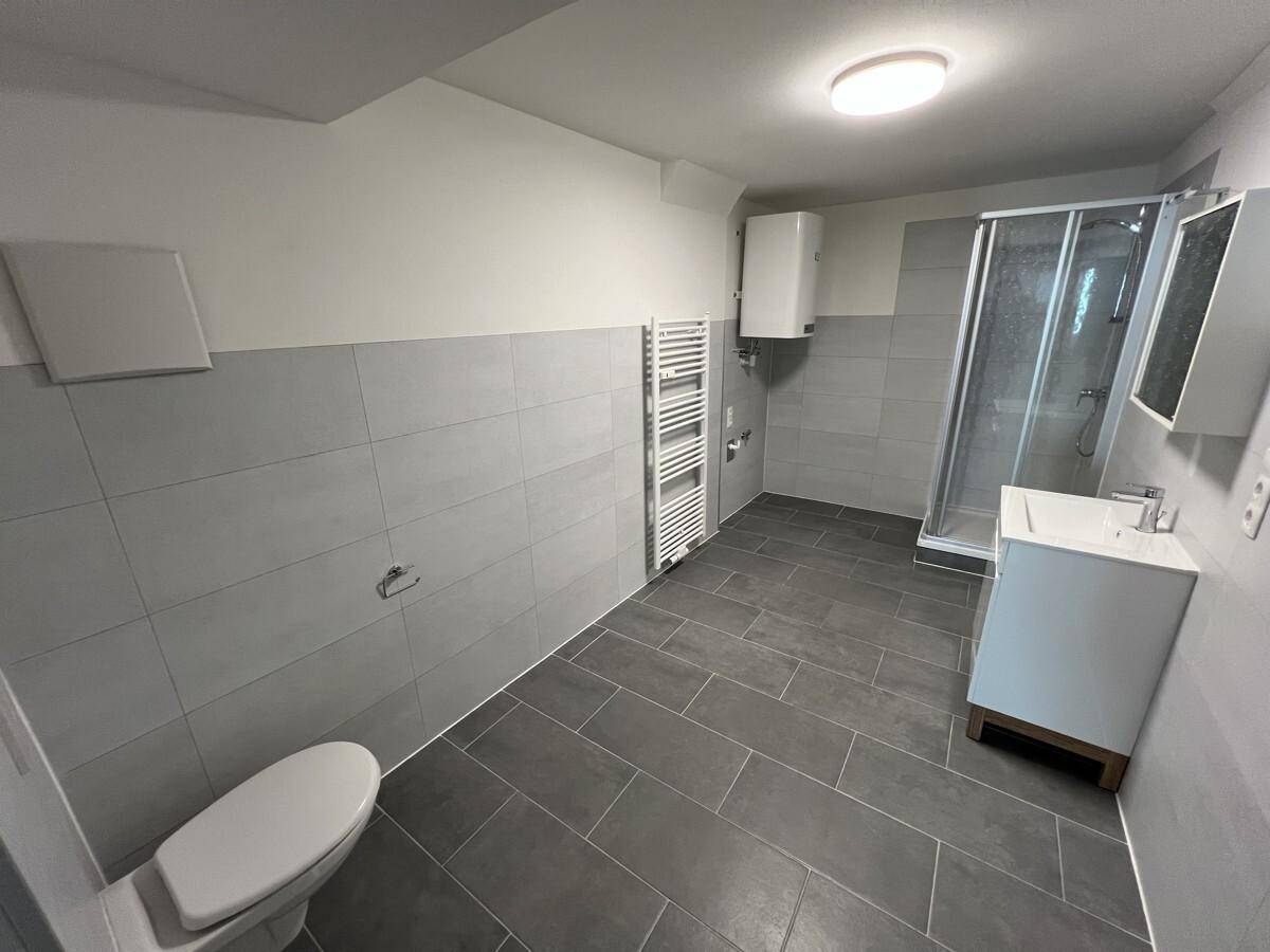 Helles Badezimmer mit grauen Fliesen, Dusche, WC und großem Warmwasserboiler.