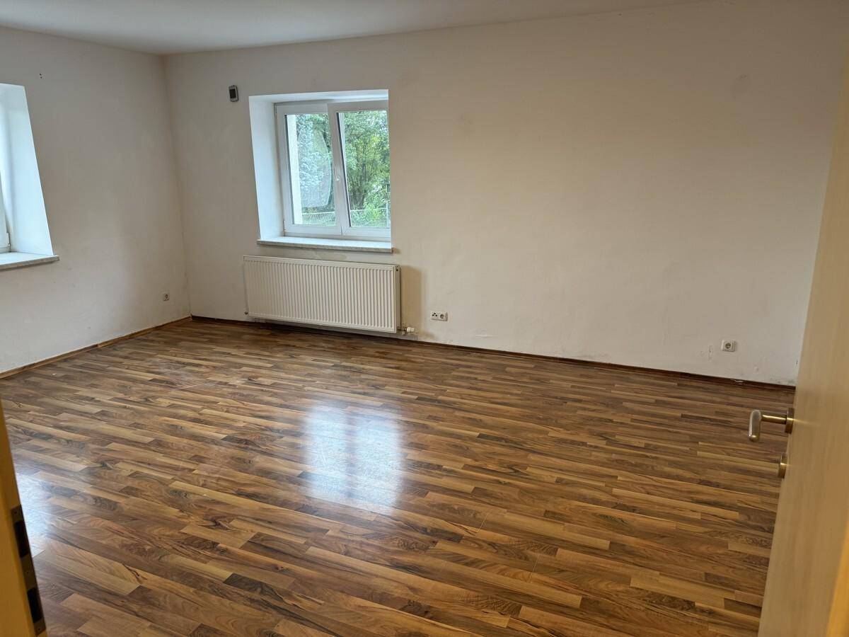 Geräumiges, leeres Zimmer mit Laminatboden und Blick ins Grüne durch das Fenster.