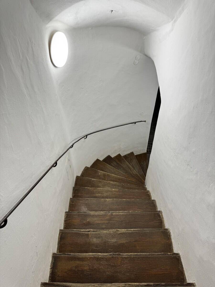 Historische Holztreppe mit Handlauf in einem engen, gewölbten Treppenhaus.