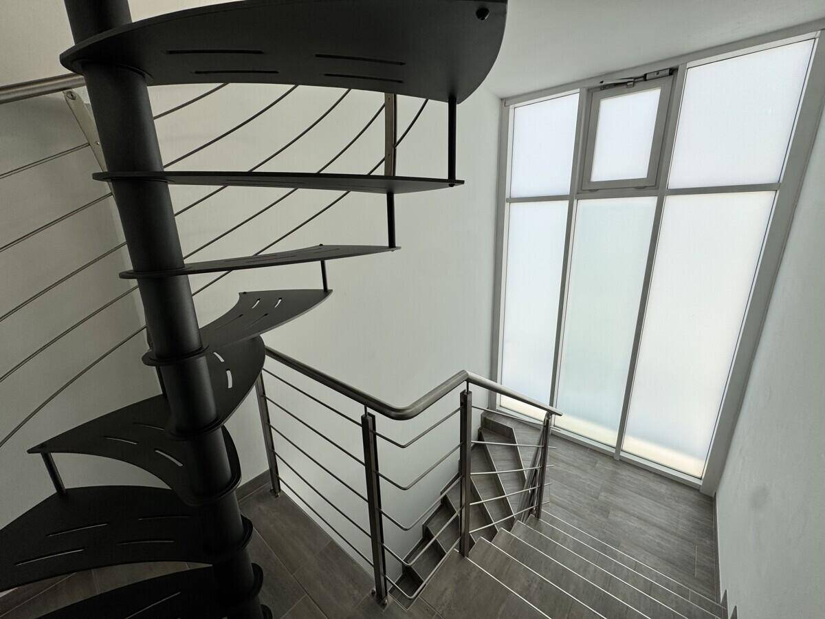 Moderne Wendeltreppe aus Metall mit Edelstahlgeländer und großen Fenstern, die den Raum erhellen.