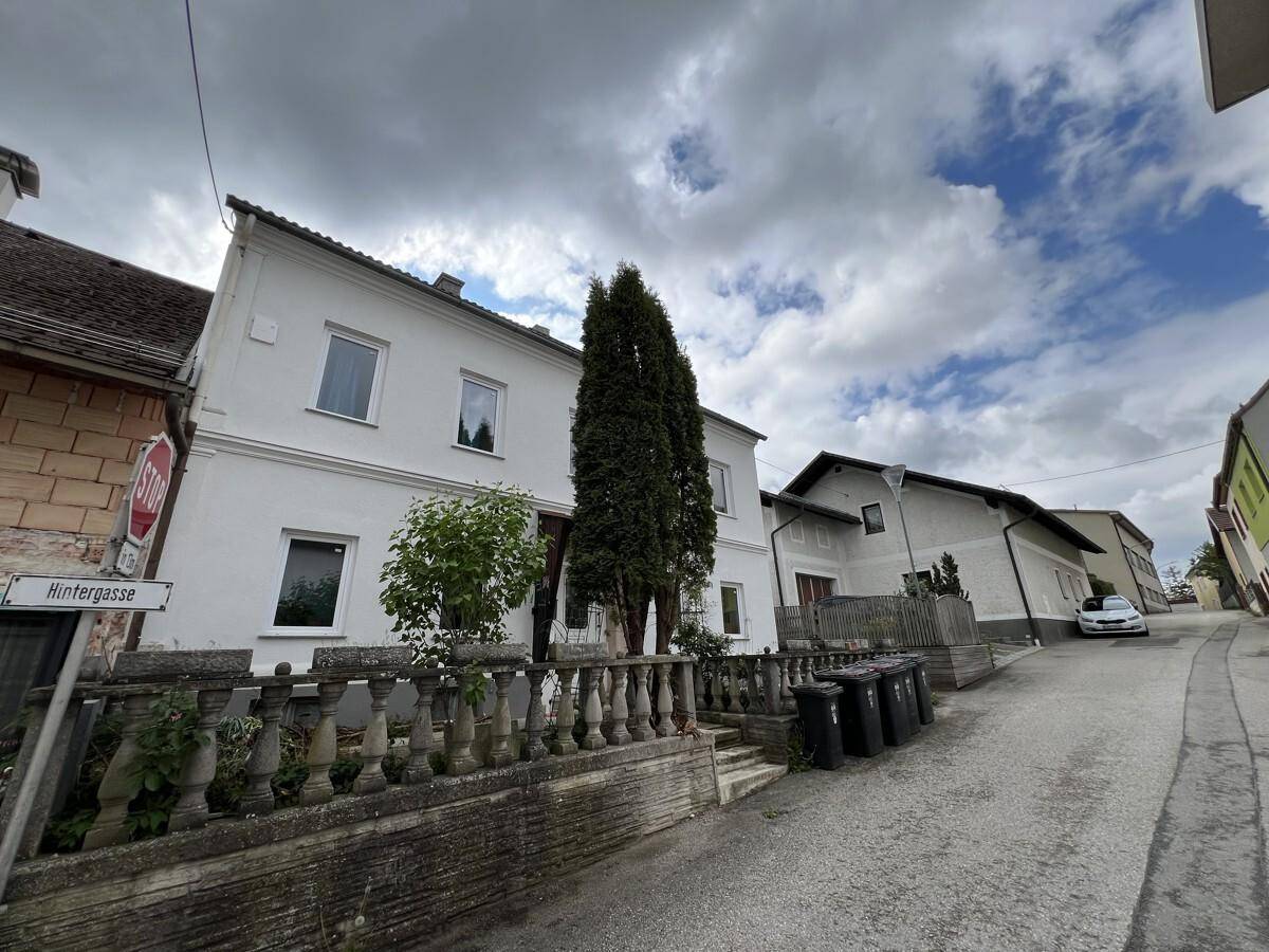 Ansicht des Wohnhauses an einer leicht ansteigenden Straße mit Mülltonnen und Nachbargebäuden unter bewölktem Himmel.