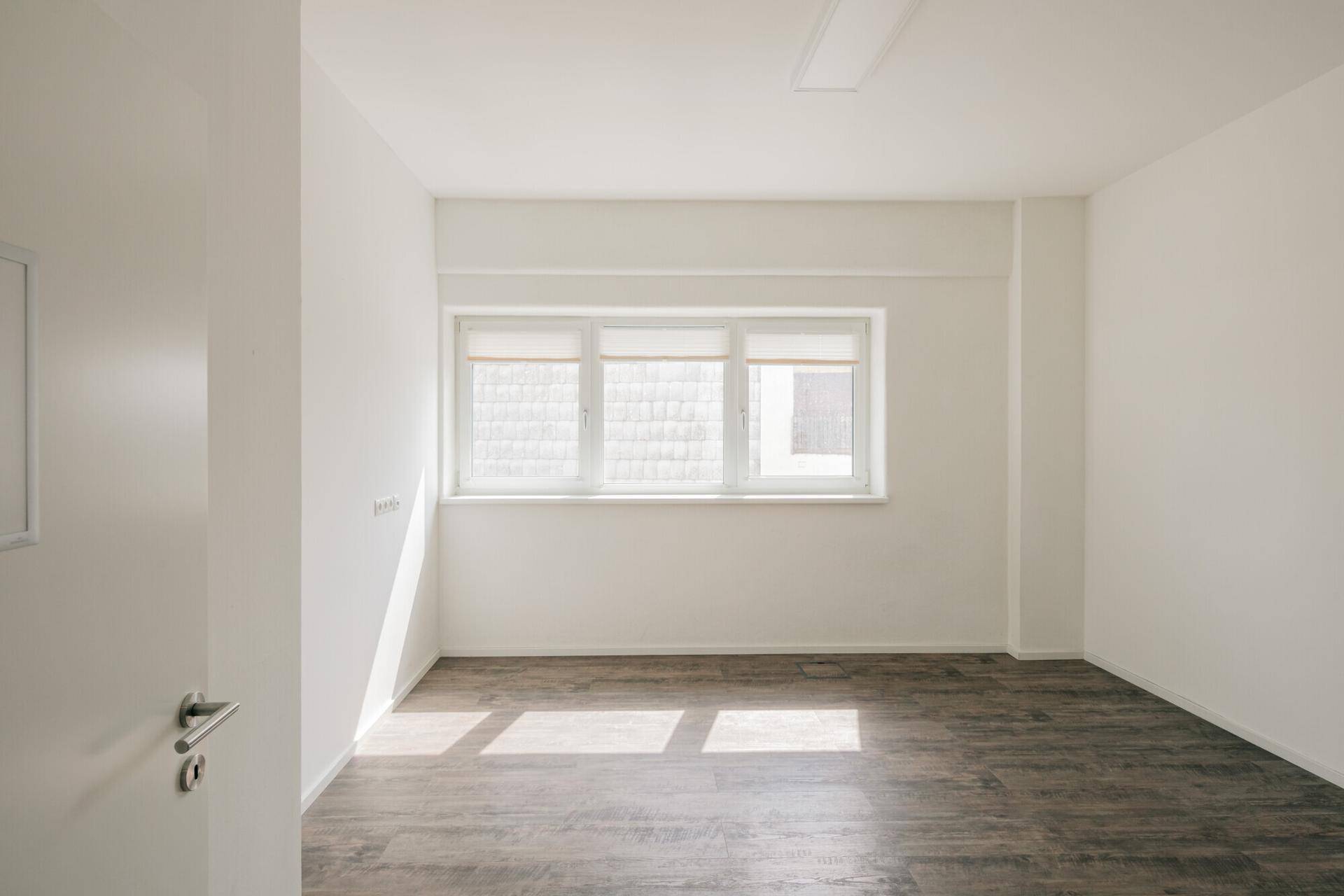 Heller Büroraum mit großen Fenstern, die viel Tageslicht hereinlassen, und dunklem Holzoptik-Boden.