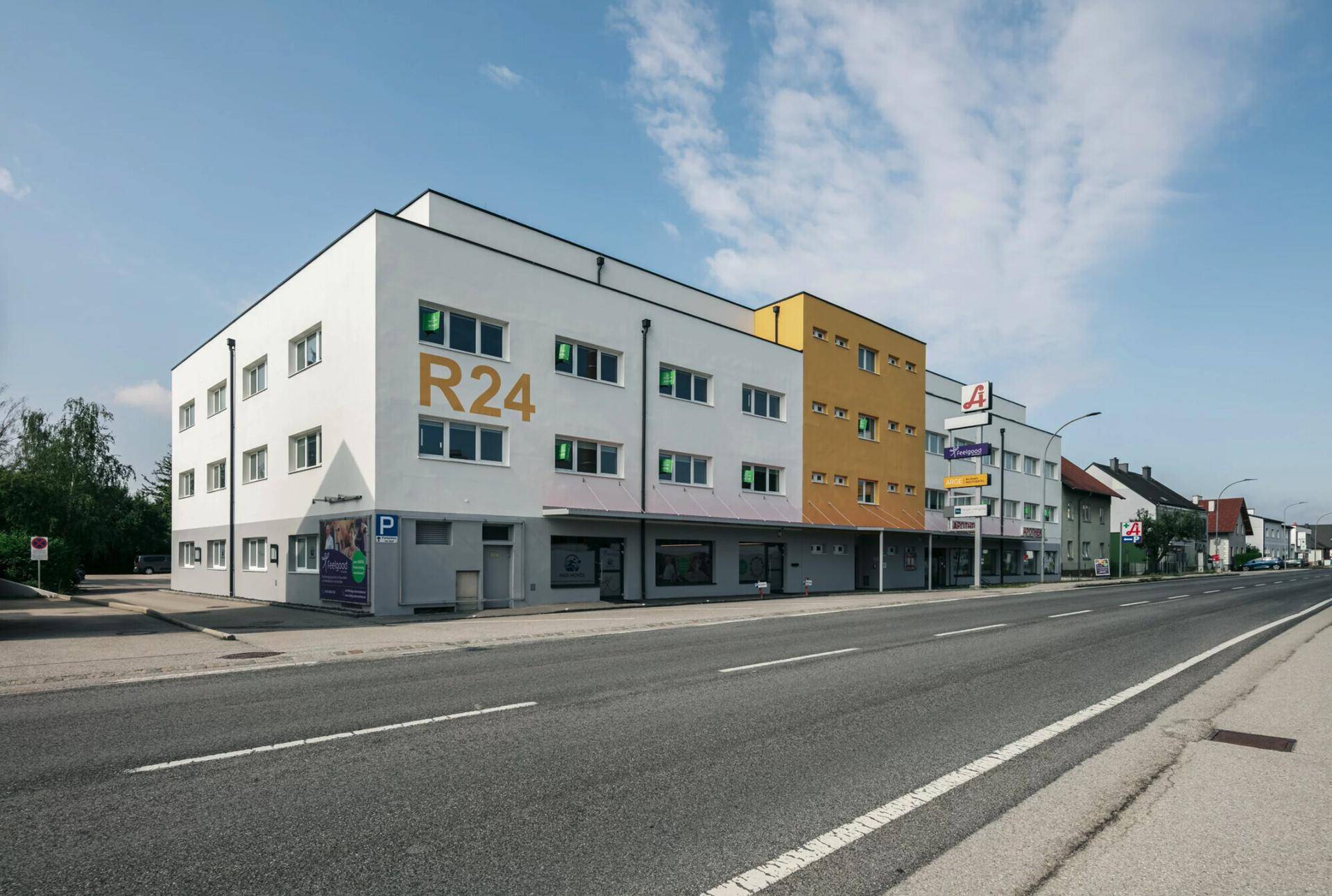 Modernes Gebäude mit weißer und gelber Fassade, Geschäften im Erdgeschoss und Parkplätzen davor.