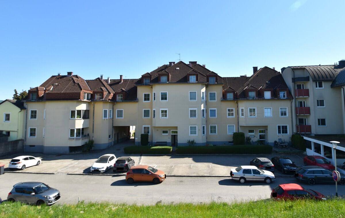 Mehrfamilienhaus mit Parkplätzen vor der Fassade unter blauem Himmel, umgeben von weiteren Gebäuden.