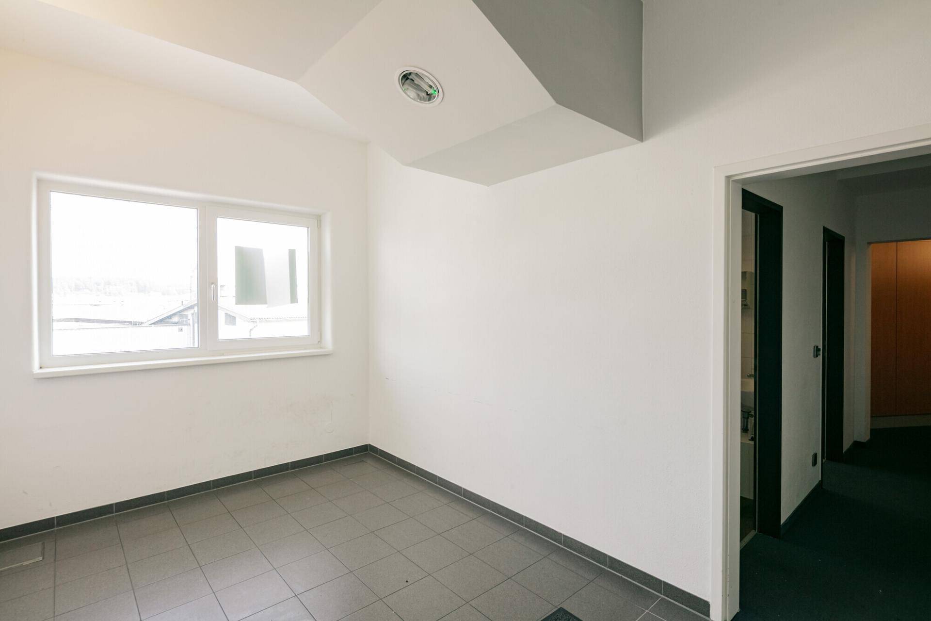Kleiner Raum mit grauem Fliesenboden, weißen Wänden und einem Fenster, ideal als Abstellraum oder kleines Büro.