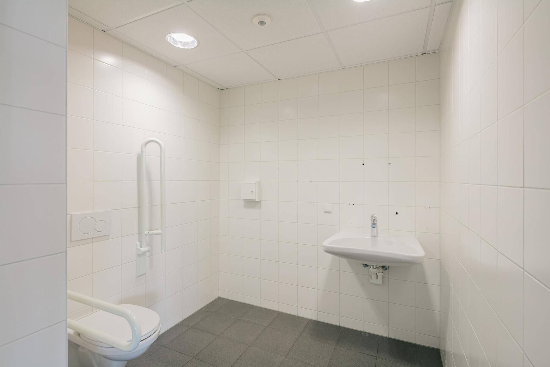 Barrierefreies Badezimmer mit weißen Fliesen, Haltegriffen und einem wandhängenden Waschbecken.