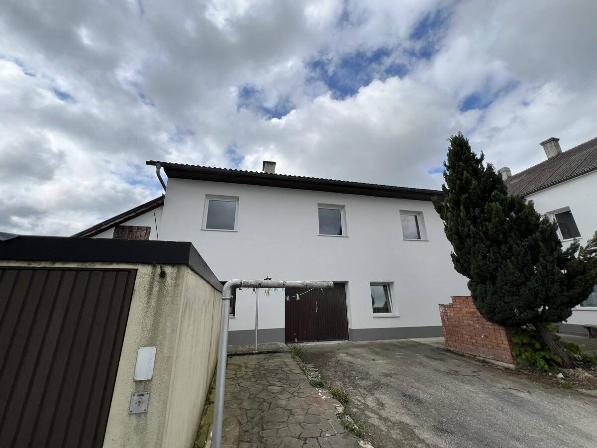 Rückansicht des Wohnhauses mit Garagentor und Zufahrt unter bewölktem Himmel, umgeben von Bäumen.