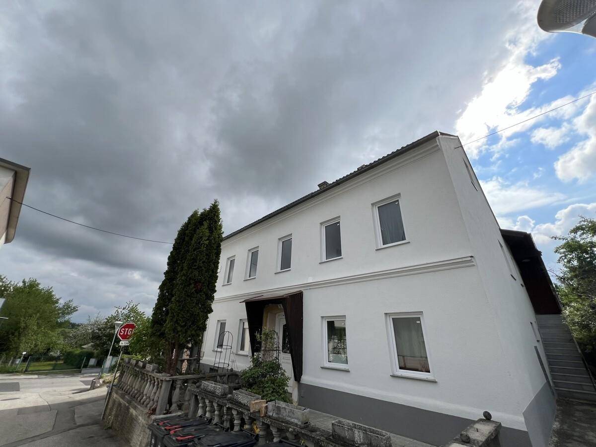 Weißes Wohnhaus von der Seite mit Blick auf die Straße und bewölkten Himmel, umgeben von Grün.