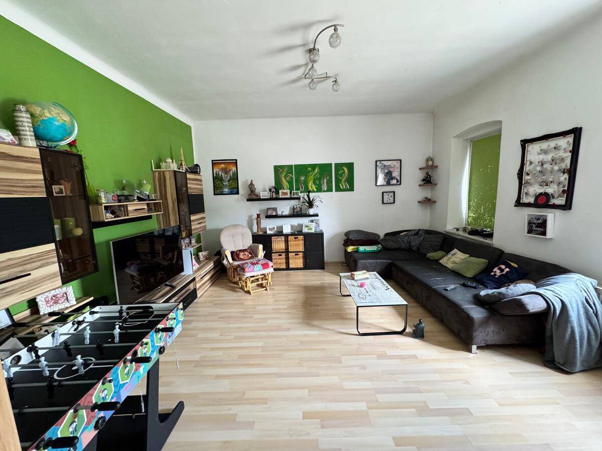 Geräumiges Wohnzimmer mit hellem Laminatboden, grüner Akzentwand und großem Sofa.