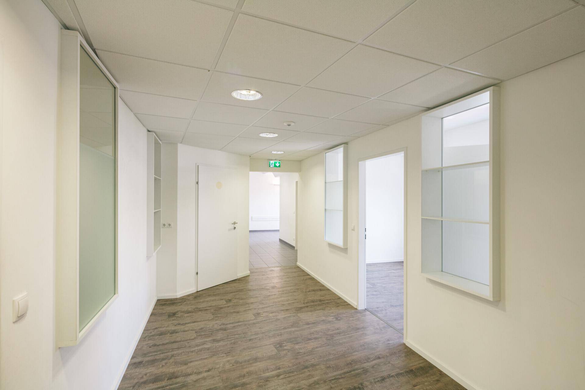 Geräumiger Flur mit Holzoptik-Boden, weißen Wänden und Deckenleuchten, der zu Büros führt.