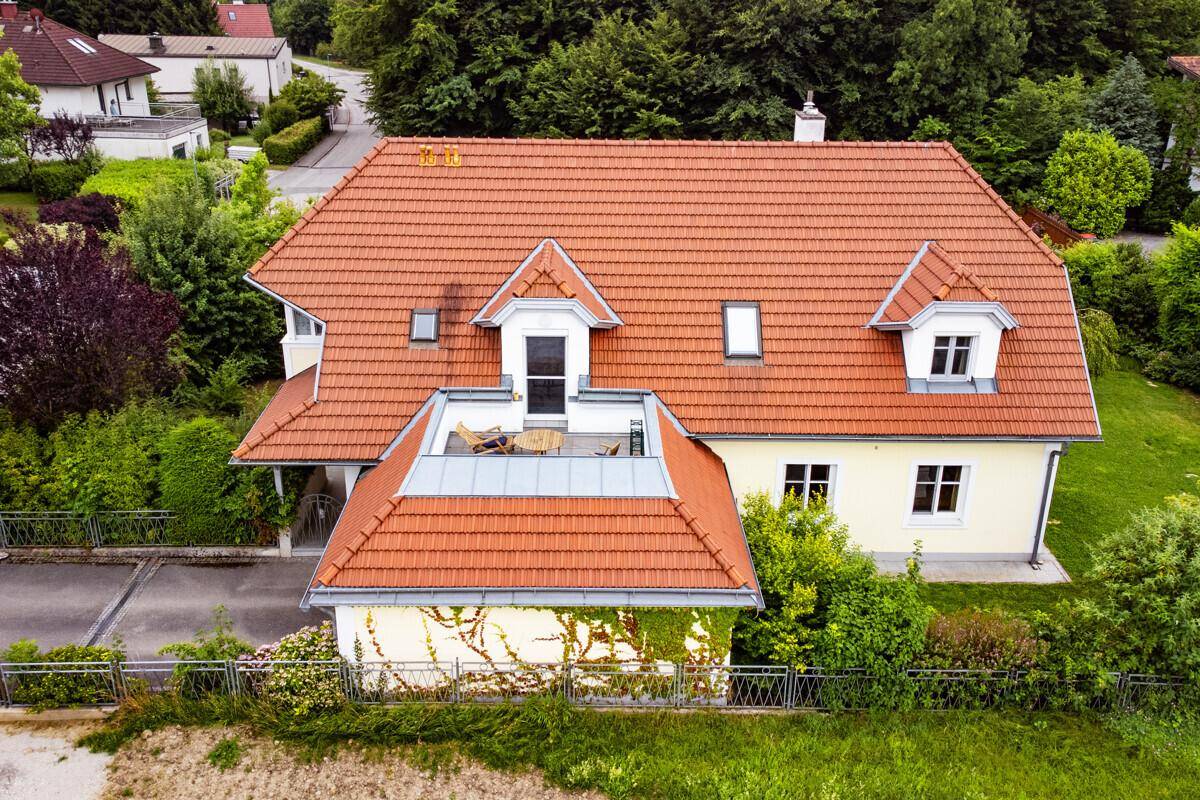 Rückansicht des Hauses mit Terrasse auf dem Dach und rotem Ziegeldach, umgeben von viel Grün.