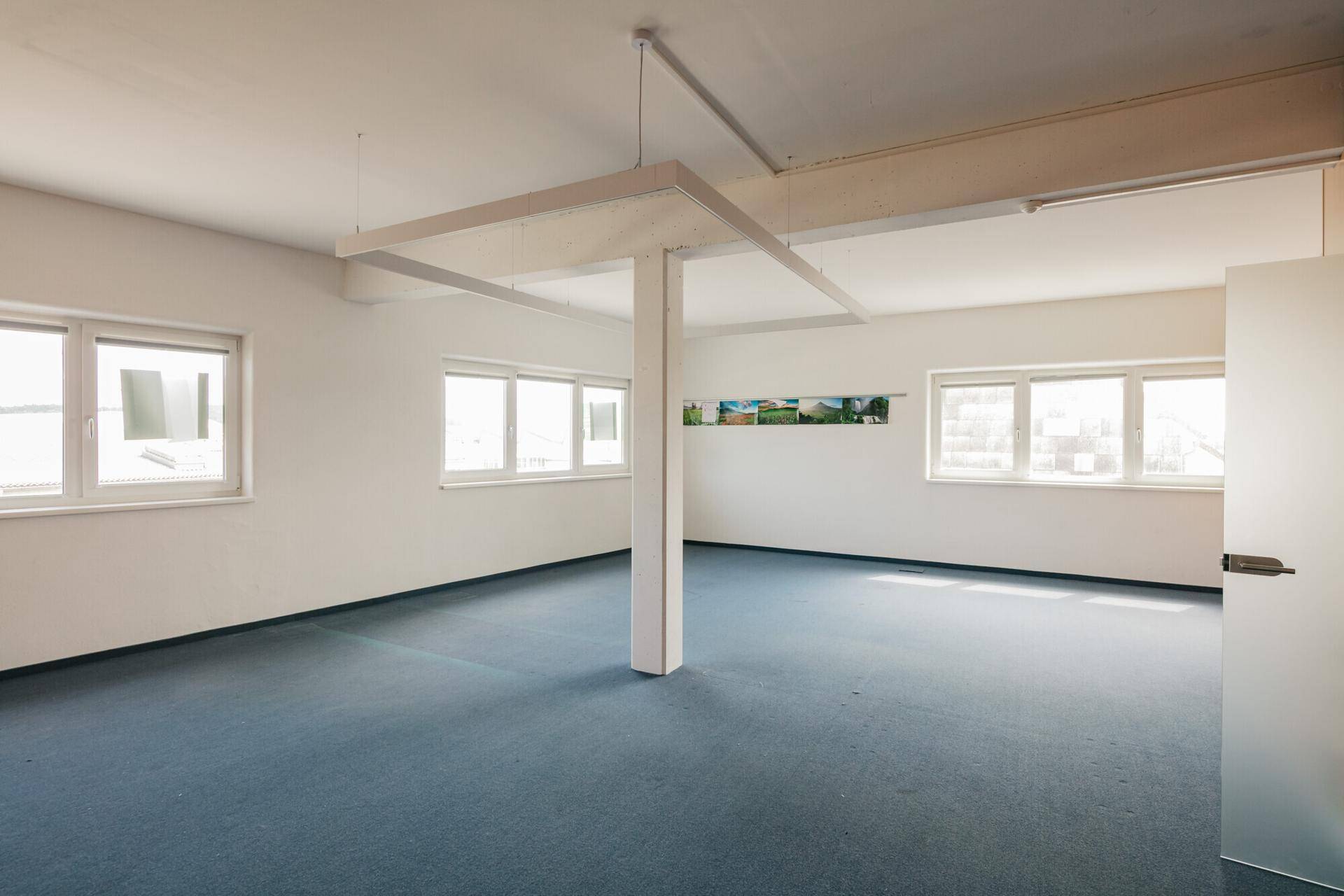 Geräumiges, leeres Büro mit blauem Teppichboden, weißen Wänden und vielen Fenstern für natürliches Licht.