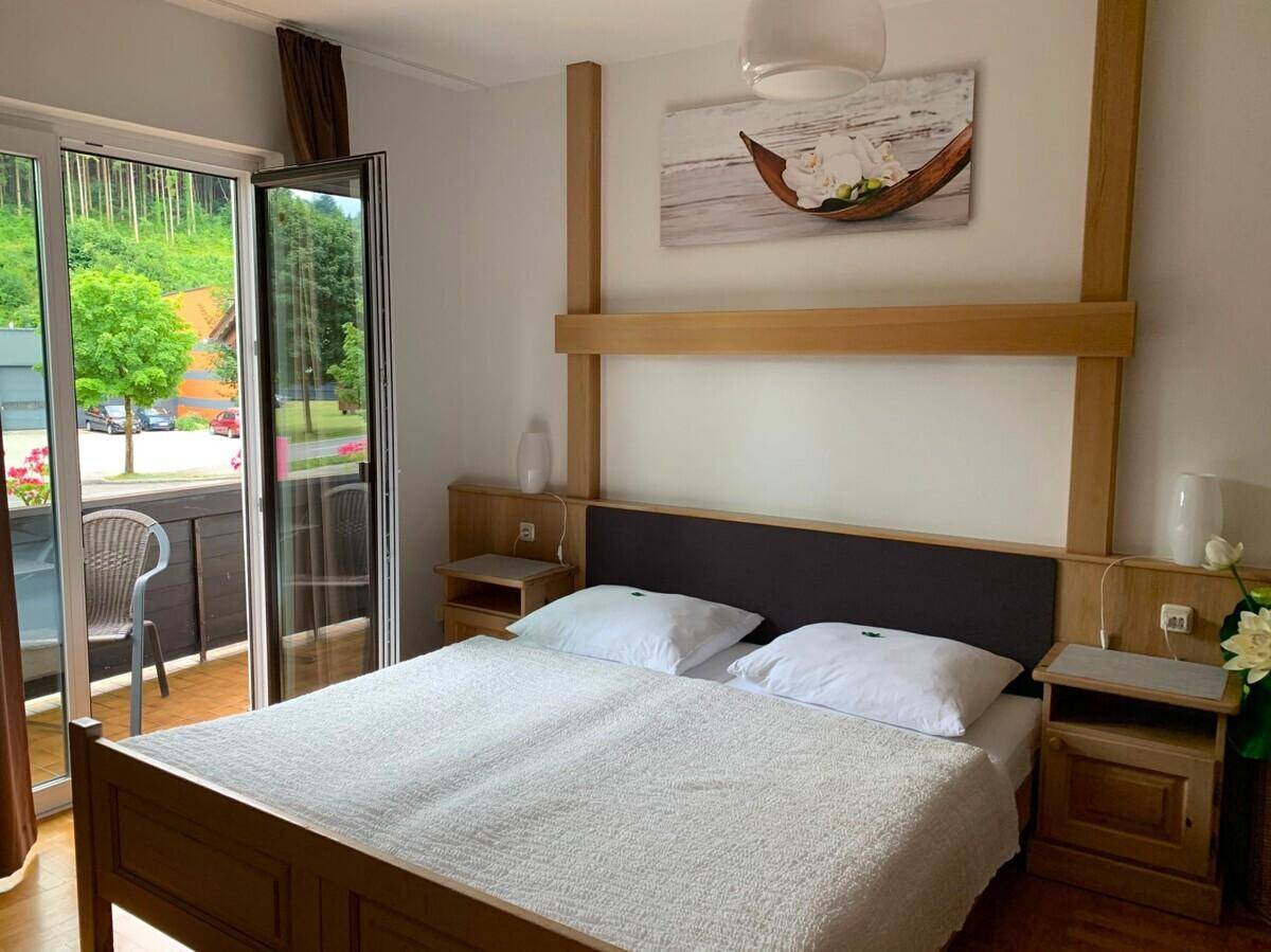 Helles Schlafzimmer mit Holzbett, Balkonzugang und Blick auf die bewaldete Umgebung.