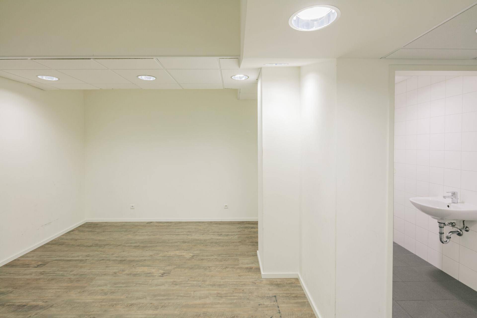 Heller Büroraum mit Holzoptik-Boden und Blick auf einen angrenzenden Sanitärbereich mit Waschbecken.