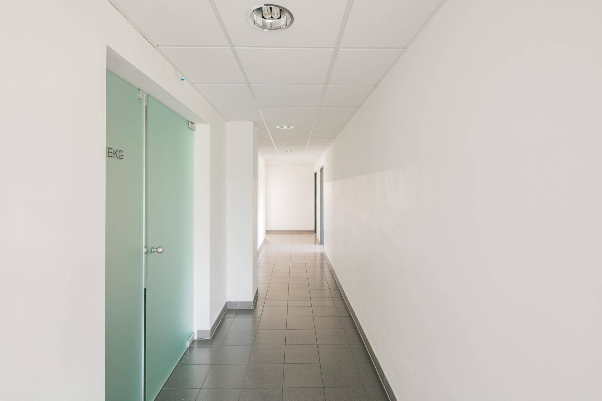 Heller Flur mit grauen Fliesen, abgehängter Decke und modernen Milchglastüren zu Behandlungsräumen.