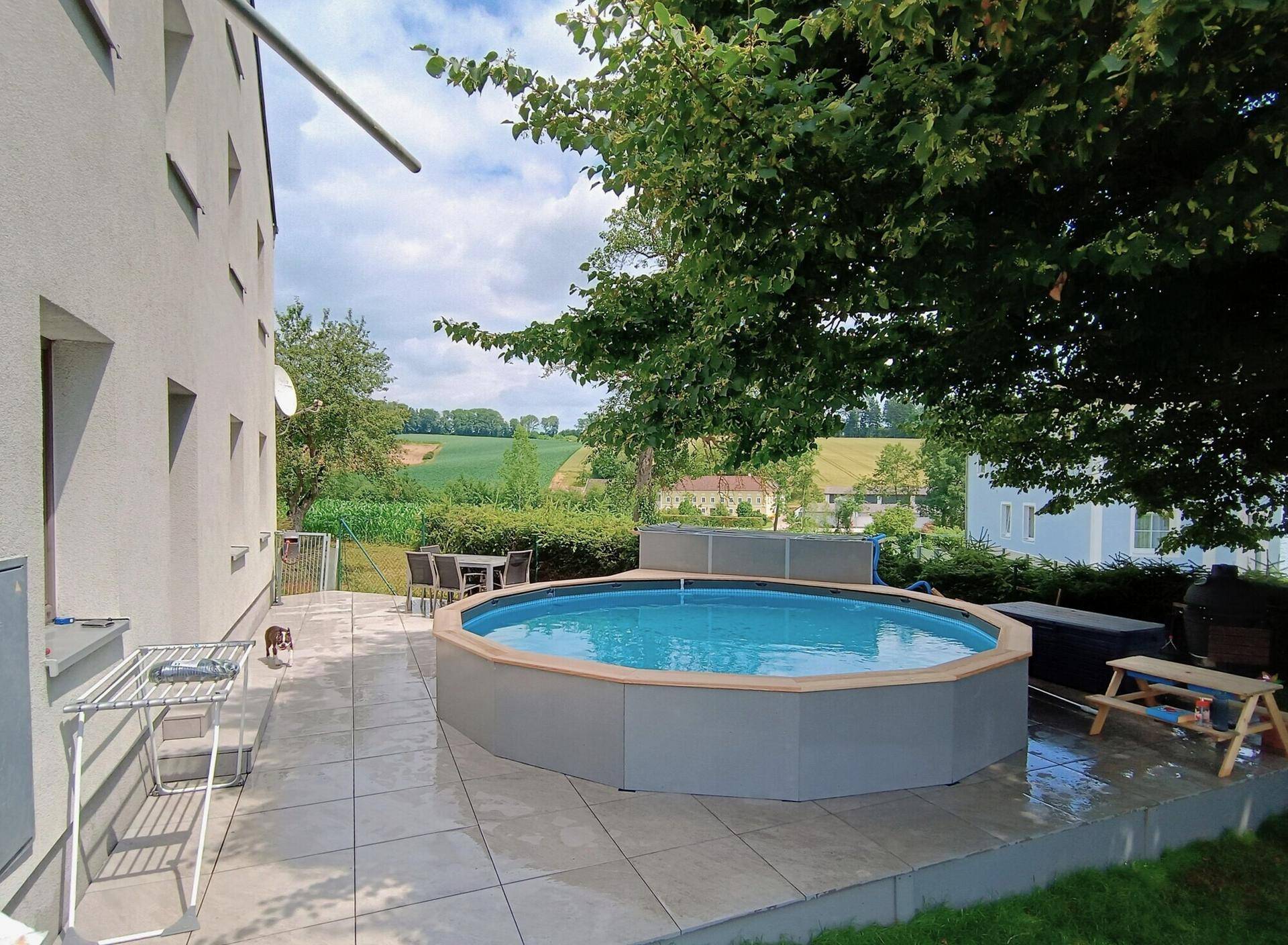 Sonnenterrasse mit Pool und Sitzgelegenheiten, umgeben von Grünflächen und Bäumen.