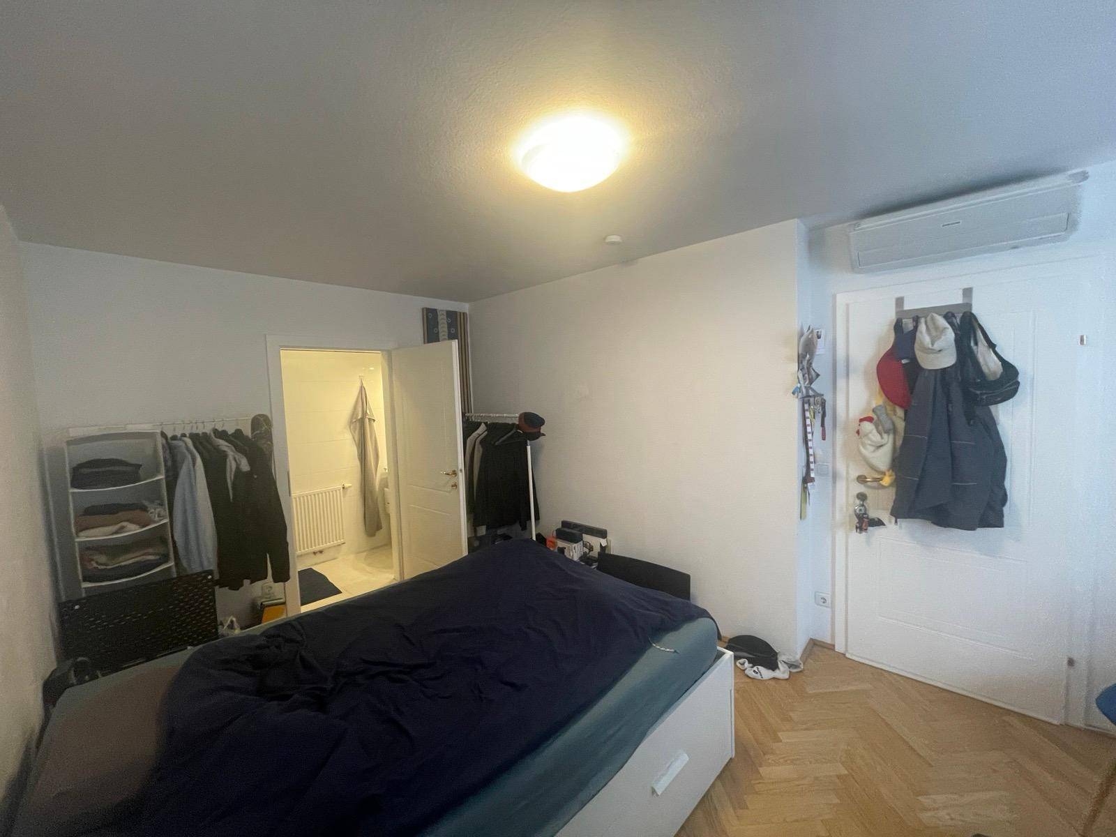 Schlafzimmer mit Parkettboden, Bett, Kleiderstange und Tür zum angrenzenden Badezimmer.