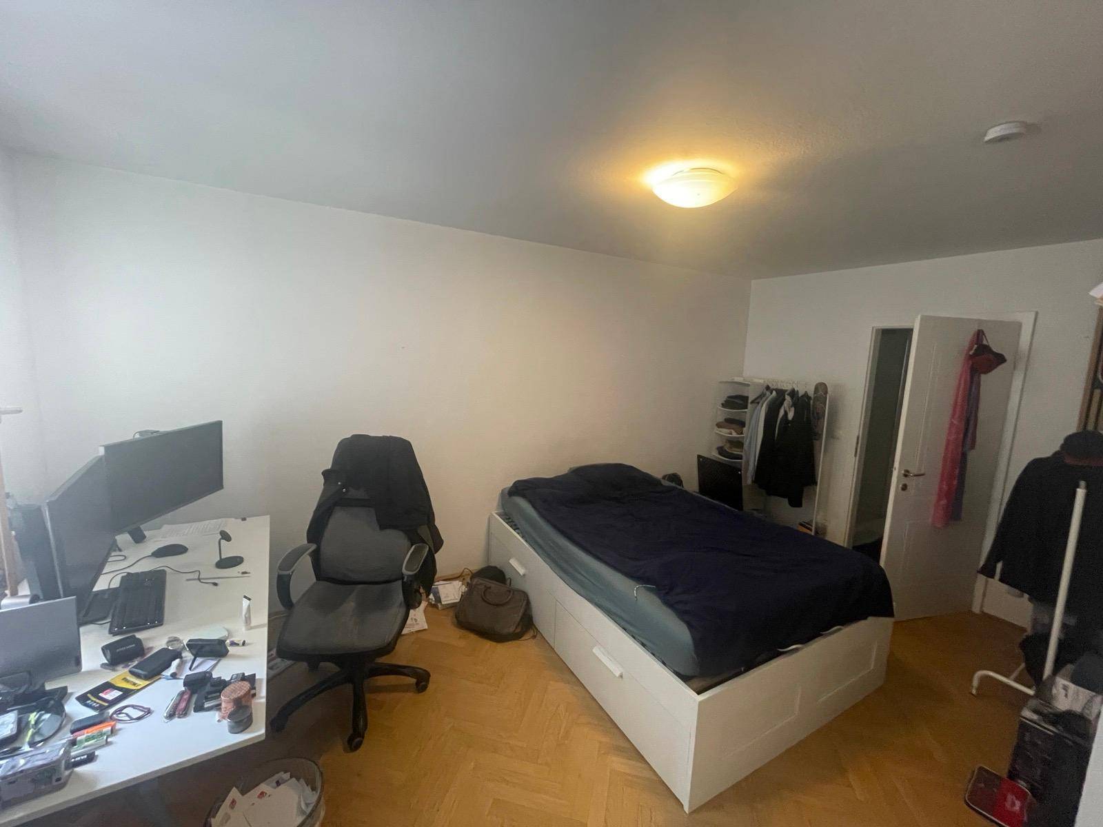 Helles Schlafzimmer mit Parkettboden, Einzelbett, Schreibtisch und Kleiderstange.
