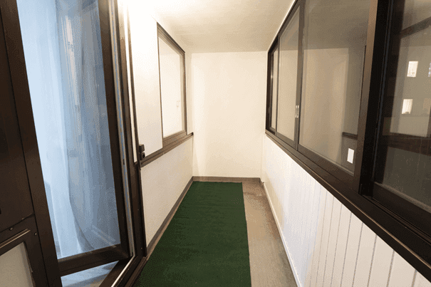 Schmale, verglaste Loggia mit dunklen Fensterrahmen und grünem Teppichboden.
