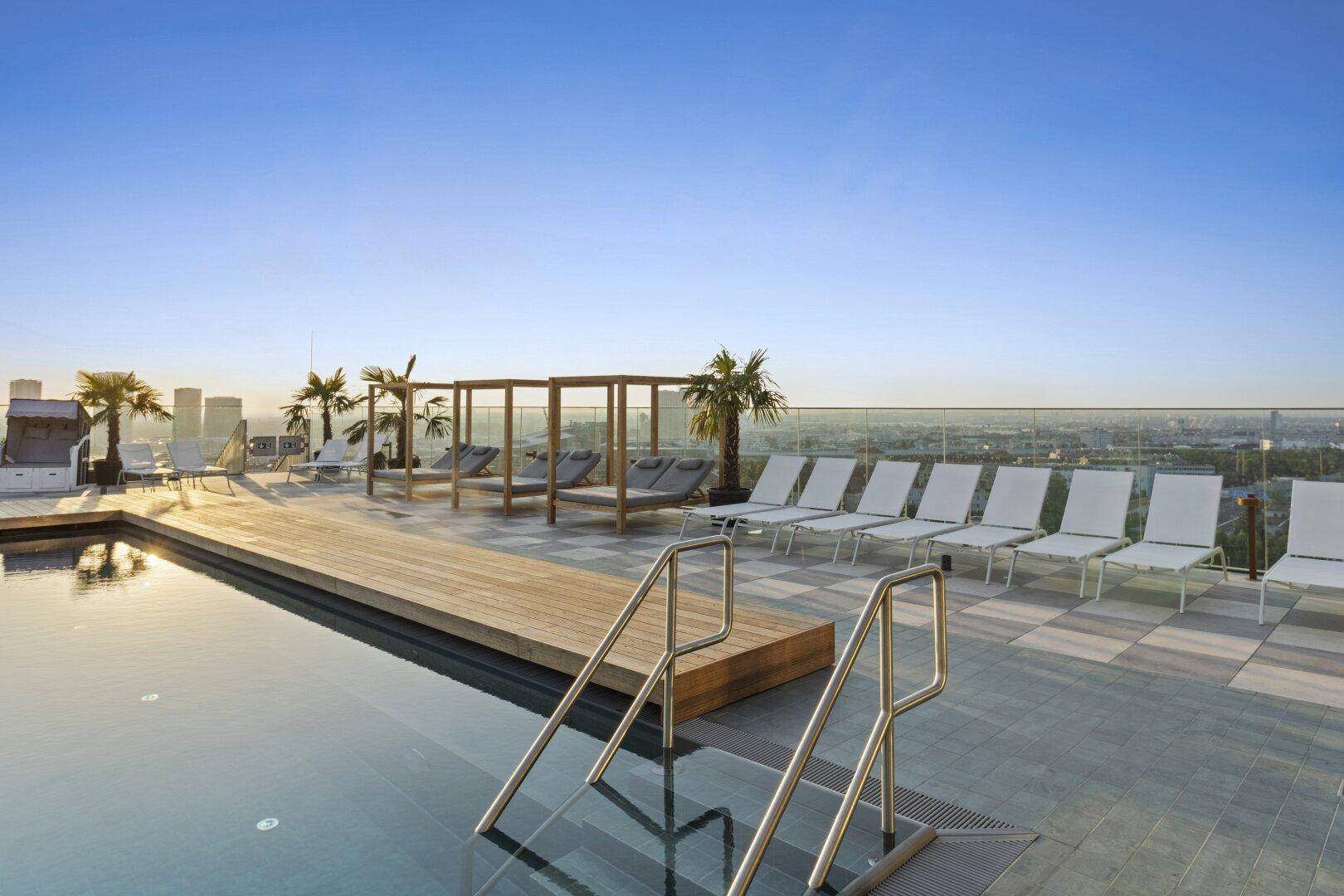 Exklusiver Dachterrassen-Poolbereich mit Liegestühlen, Palmen und weitem Stadtblick bei Sonnenuntergang.