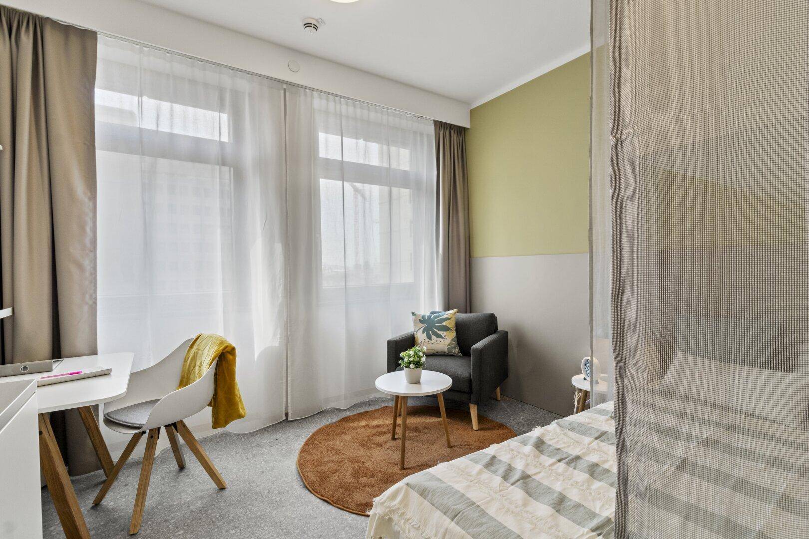 Heller Studio-Wohnraum mit Schlafbereich, gemütlichem Sessel und Schreibtisch am großen Fenster.
