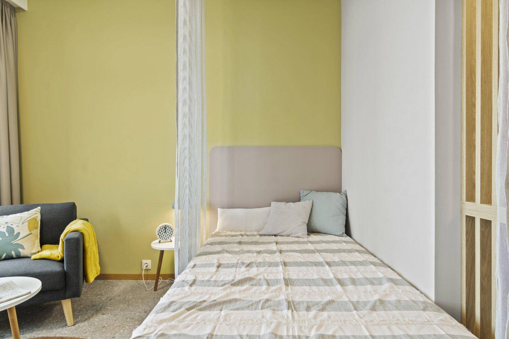 Schlafbereich in einem Studio-Apartment mit Vorhang als Raumteiler und gelber Wand.