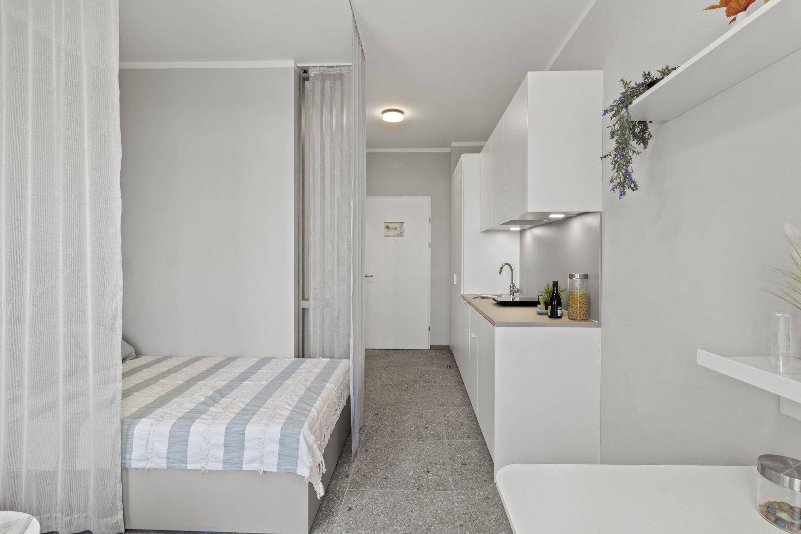 Effiziente Küchenzeile mit weißen Fronten und angrenzendem Schlafbereich in einem Studio-Apartment.