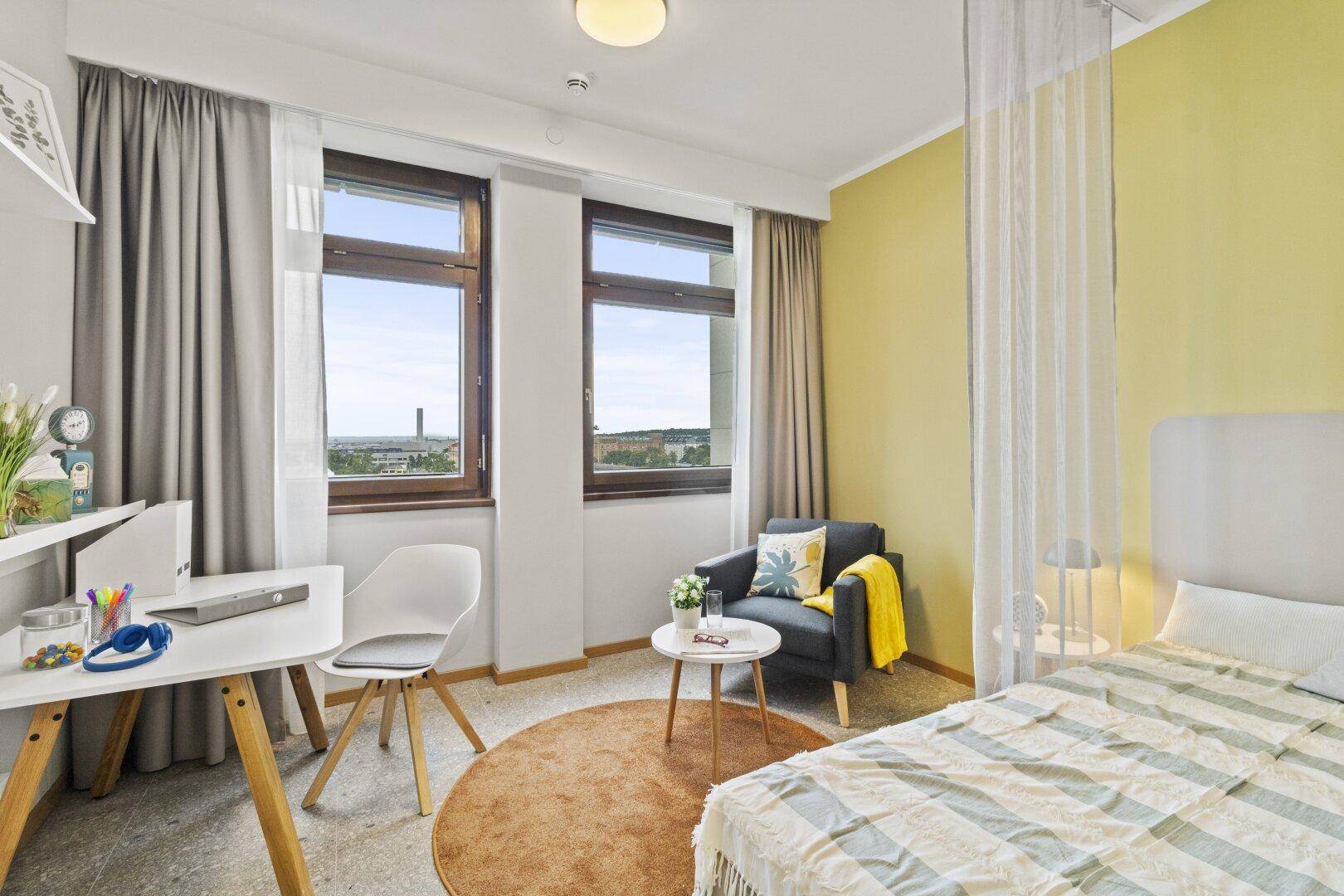 Helles Schlafzimmer in einem Studio-Apartment mit Schreibtisch, Sessel und großen Fenstern.